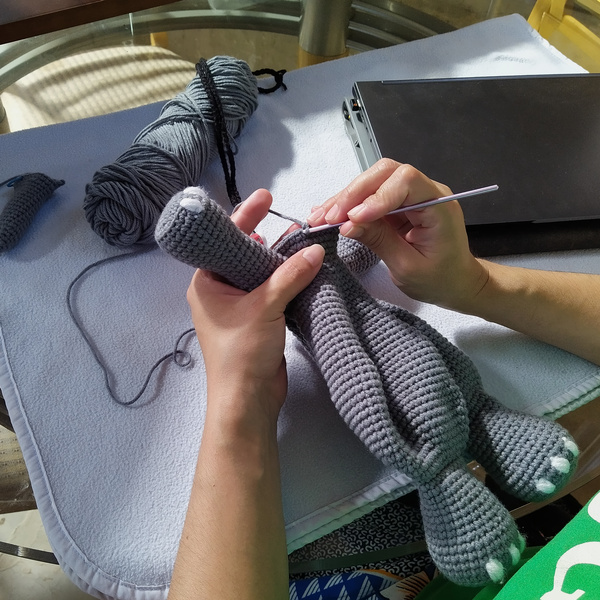 A view from over the left shoulder of a woman crocheting the neck area of an elephant lovey. A closed laptop is visible on the right, along with the yarn skein being used.