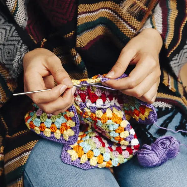 Hands crocheting a colorful granny square blanket with purple yarn.
